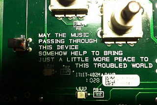 A circuit-board with text inscribed upon it, saying “may the music passing through this device somehow help to bring just a little more peace to this troubled world”.