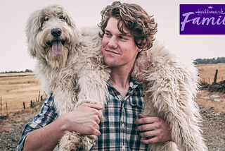 Young man with a plaid shirt on a country lane carrying a shaggy dog with the Hallmark Channel icon in the upper right corner.