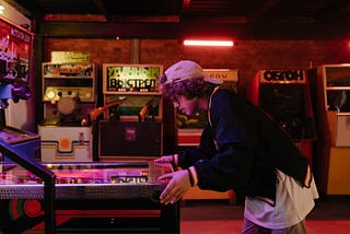 A boy in a white cap and black jacket leans over a pinball machine in a tattered arcade dimly lit by orange neon strips.