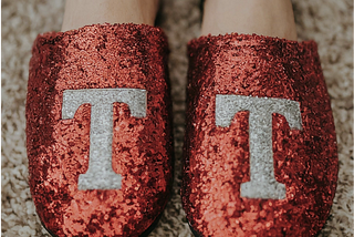 Feet wearing sparkly red slippers emblazoned with the letter T in silver