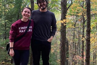 a short white transmasc human in a burgundy “KINDNESS MATTERS” sweatshirt in the woods of Vermont in fall 2020, next to John Maher, a white person with chin length brown hair and a mustache and beard, wearing a dark grey sweater and black pants
