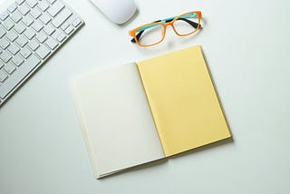 A picture of a keyboard, a computer mouse, eye glasses, and an open note showing empty pages.