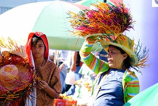 A woman wearing a colorful outfit holds a handmade hat adorned with vibrant strips of straw, while a young boy with a serious expression stands beside her, holding additional hats for sale. The scene is set under a bright, sunny day at a bustling festival, with colorful umbrellas in the background providing shade for vendors and attendees. The image captures a mix of livelihood and tradition amidst the celebratory atmosphere.
