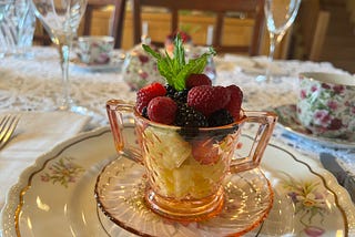 A table set with a lace tablecloth and floral dishes along with a pink glass teacup filled with fruit salad and garnished with fresh mint.