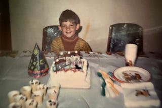 Boy sitting at at table with a birthday cake and party decorations.