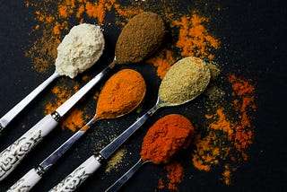 Five metal spoons carrying various colorful spices on a black countertop.
