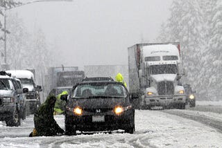 A person is kneeling in the middle of the road during a snowstorm to look at the tires of the car. There are skid marks and heavy traffic surrounding the car.