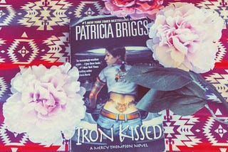 Three flowers, two pink and one red, bordering the book “Iron Kissed” by Patricia Briggs. Photo Credit, Zirrina Maxwell. February 9, 2024.