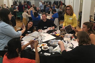 A group of people standing and sitting around a round table. The table is covered with newspapers and magazines and sweets. They are talking and showing each other things that they have cut out. In the background more tables are visible.