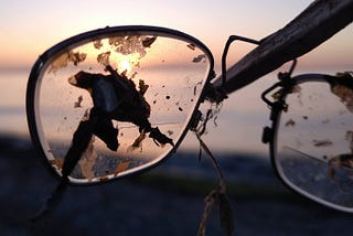 Cracked lens in an eye-glass frame held up by a stick with sun setting on the ocean in the background.
