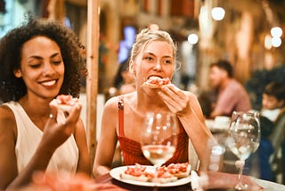 A blonde woman and a black woman sit eating pizza with wine glasses in front of them.