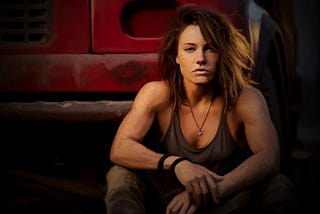 Digital painting with a warm palette of a young butch woman with long brown hair and freckles in a tank top sitting in front of an old red truck.