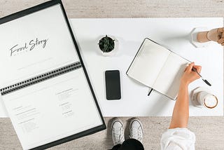 Flat lay overhead photograph of an office desk with copywriter writing in notebook, phone and brand book. Image by Food Story Media digital agency in London.