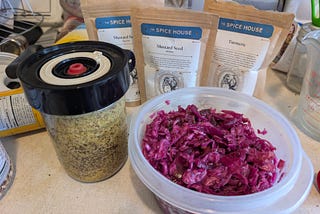 On the left, a glass jar with a fermentation lid (the lid has a valve in the center to release excess CO2 gas) containing yellow and brown mustard seeds and other ingredients. On the right, in a plastic container, reddish-purple sauerkraut made from red cabbage. In the background, three spice pouches from The Spice House — turmeric and two types of mustard seed.