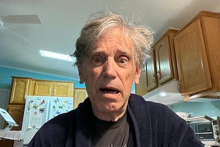 A surprised-looking man with disheveled gray hair wearing a dark robe and a T-shirt, standing in a kitchen. The background shows wooden cabinets, a refrigerator with colorful magnets, and a ceiling fan above. The expression on his face suggests shock or confusion.