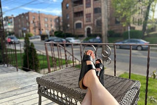 A street, a porch, an outdoor ottoman, and Amy’s legs wearing black Birkenstocks. The sun shines and the first perspective suggests calm.