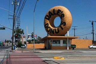 A corner donut shop is dwarfed by a gigantic donut statue mounted on the roof. The donut is painted with bold-face text “Dale’s Donuts.”
