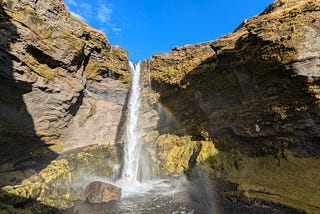 A waterfall cascades over a rocky cliff with a rainbow in front