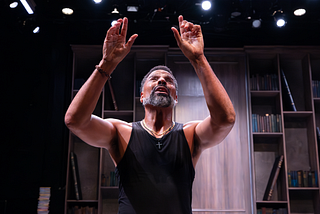 A man in a black tank top stands in front of a wood-walled library under stage lights and speaks with his arms raised.