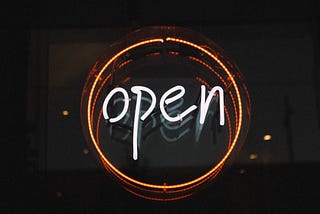 Neon “open” sign with white text, surrounded by orange ring, on a black background. Image has dim reflection.