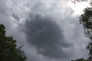 A dark cloud against lighter clouds, framed on three sides by tree branches.