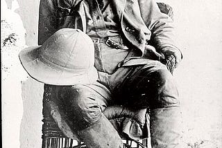 Photograph of Theodore Roosevelt sitting in a chair in expedition attire. Hat on his lap.