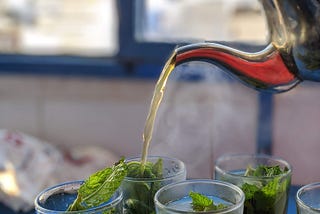 Photo of a metal teapot pouring hot water into glasses of fresh mint.
