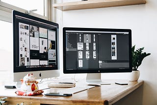 Two silver and black iMacs displaying design workflows sit on a wooden desk against a sunlit window