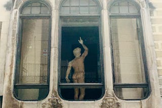 An outside window view of a statue of a naked man, from behind. He is looking up and pointing.