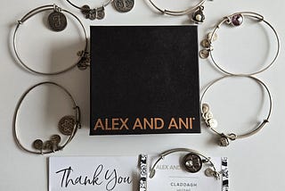Black 3 1/2" square jewelry box with Alex and Ani on it in gold capital letters, surrounded by silver wire bracelets bearing charms featuring a script letter F, a Jewish star, a Claddagh, a purple gemstone, a weathered silver flower with a pearl in the middle, and a tree of life