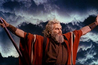 Color movie still of Charlton Heston as Moses in The Ten Commandments, 1956, with arms outstretched against a dark sky, parting the Red Sea.