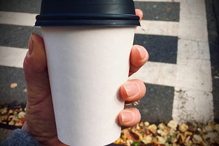 A plain white coffee cup with a black lid is grasped by a hand, taking up almost the whole frame. The background shows a crosswalk with a pile of leaves gathered at the road’s edge.