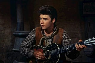 Cropped colour screenshot of singer Ricky Nelson holding a guitar whilst singing taken from the trailer for the Howard Hawks 1959 film “Rio Bravo”.