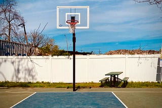 Image shows a lone basketball hoop in front of a white fence, with a blue sky behind it.