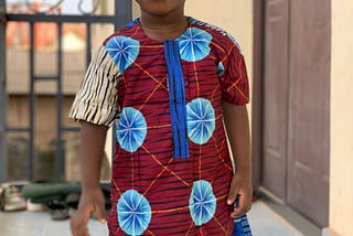 Young brown boy wearing African print and standing in front of a brown door.