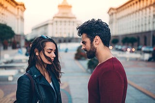 A woman and a man stand together in a downtown area, looking sad. The woman is dark-haired, in a black leather jacket, looking down in regret. The man is dark-haired as well with a short beard, and a long sleeved maroon shirt, looking at her in hurt.