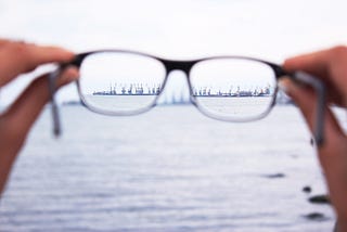 Imagem fotográfica de um par de óculos de grau sendo segurado por uma pessoa, focando guindastes em um porto do outro lado do mar. O céu está nublado, criando uma paisagem predominantemente cinza.”