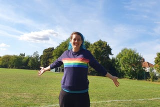 A photo of the author standing in a field, their arms are open to the side and they are smiling, the sky is blue overhead and it’s a sunny day, they are wearing a navy blue pumping jumper with a rainbow stripe across the chest