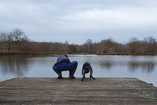 A woman and her dog, both crouched on a wooden dock over a lake, both leaning slightly to the left in sink.