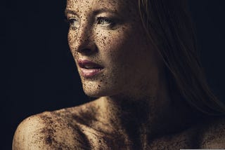 A woman’s face and shoulder against a black background. Her skin is covered with dirt.