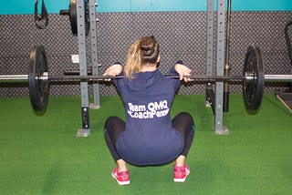 Image shows coach pennie demonstrating a full-range front squat. They are wearing pink crossfit nanos, black leggings and a blue shirt with #coachpennie printed on the back.