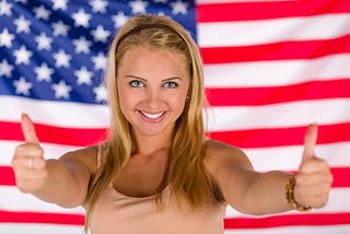 A smiling blonde-haired, blue-eyed white woman gives us two thumbs us while standing in front of an American flag.