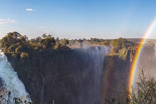 A set of three massive waterfalls surrounded by lush plant life and large trees basking in the sunshine. Water spray causes a vivid rainbow to appear against the cliffs and the pristine blue sky.