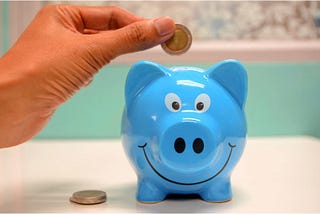 A hand dropping a coin into a piggybank; you can also save money as a writer by getting free writing lessons