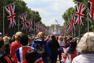 A gaggle of bored british citizens stand in a makeshift queue to nowhere while an endless stream of Union Jacks line an arboreus corridor forcing the crowd’s attention toward the gilded idol that stands before them in the distance.