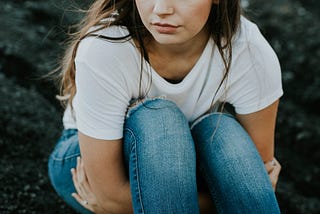 A girl with social anxiety disorder in a white T-shirt and jeans sitting down with her knees to her chest looking sad.