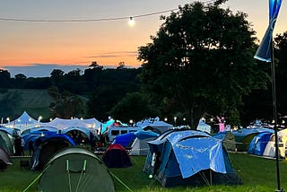 Tents in a field at dusk. A lightbulb glows on a cable above them, against the orange sky.