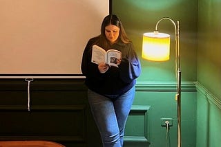 Yasmine standing up next to a lamp holding a book.