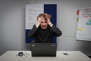 A man is sitting at a desk in front of a laptop with his hands on his head, messy hair, mouth wide open and anguished look on his face. A phone, computer mouse and a pair of reading glasses sit to the side of the laptop. There is a blue board and flip chart in the background with writing on it.
