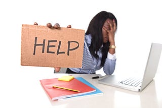 Black woman stressed out in front of her laptop holding a sign that says HELP.